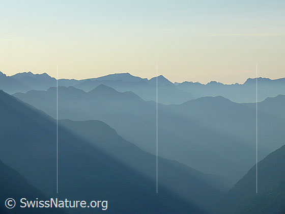 Foto: Bergketten im Morgenlicht. Die Berggipfel Vogelberg, Cima Rossa, Cima dei Cogn und der Taleinschnitt der Leventina sind in der Morgenstimmung als Silhouetten erkennbar.