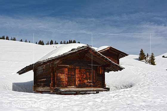 Foto: Schneelandschaft mit Alphütten und blauem Himmel, verziert mit Schleierwolken.