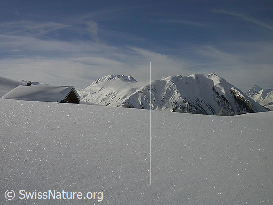 Foto: Unberührte Winterlandschaft mit eingeschneiter Alphütte. Im Hintergrund sind Berge (Bättlihorn, Breithorn) sichtbar.