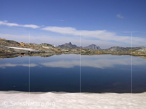 Foto: Dunkler Bergsee mm Fuss der W-Flanke des Fanellhorn: Blick über den See Richtung NNW. Spiegelung. Im Hintergund der Piz Terri. Im Vordergrund Schnee.