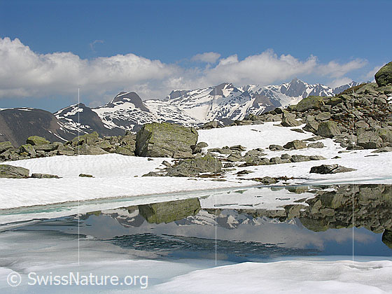 Foto: Spiegelung von Steinblock, Bergen und Wolken in Bergsee. Der Bergsee ist z.T mit Eis bedeckt.
