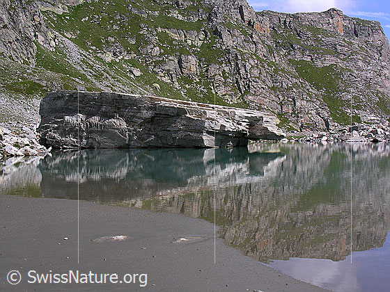 Foto: Grosser Felsblock mit Spiegelung im Mättitalsee. Das Ufer im Vordergrund ist sandig. Die Hänge im Hintergrund sind mit Gras und Fels durchsetzt.