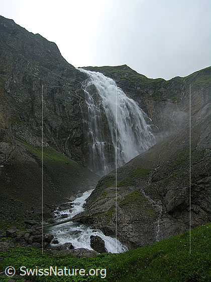 Foto: Grosser Wasserfall der Engstligenfälle. Das Wasser stürzt tosend über eine hohe Felswand in eine Schlucht und fliesst weiter unten als sprudelnder Bergbach ins Tal. Über den Bach führt ein schmaler Steg.