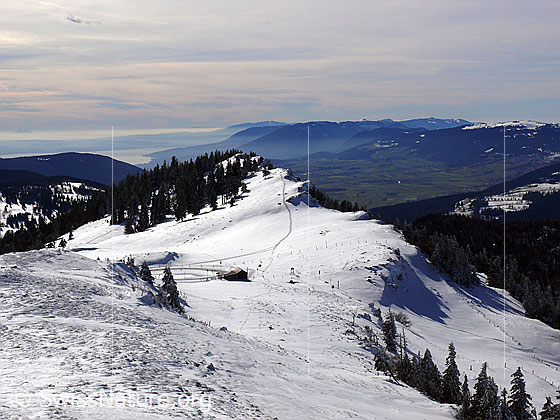 Foto: Blick über die schneebedeckte Krete des Chasseral auf den Neuenburgersee und zu den Höhenzügen des Jura mit Mont Tendre, Mont Aubert, Montagne de Boudry, Creux du Van und Mont Racine. Der Himmel ist mit Schichtwolken bedeckt.
