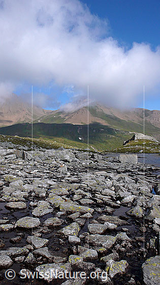 Foto: Stimmungsbild Steine, Wasser, Berge und Wolken. Blick über die Steine im seichten Wasser eines ruhigen Bergbachs zu den rötlichbraunen Hängen der gegenüber liegenden Gipfel. Darüber ziehen Quellwoken auf.