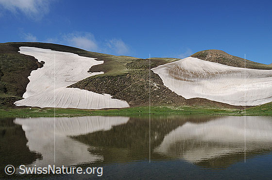 Foto: Liebliche Berglandschaft mit Schneefeldern und Bergsee mit Spiegelung.