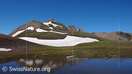 Foto: See und Berglandschaft. Karge Landschaft, Alpweiden und Schneefelder wechseln sich ab. Im Bergsee ist eine schwache Spiegelung zu sehen.