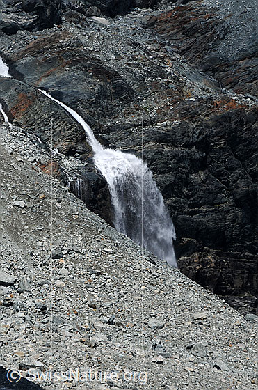 Foto: Wasserfall in karger Landschaft.
Im Fels eingefressener Bergbach und Wasserfall, welcher sich über eine Felsstufe stürzt. Die Umgebung ist karg und im Vordergrund ist ein Geröllfeld zu sehen.
