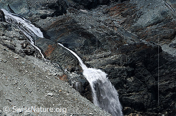 Foto: Geröll und Fels mit Wasserfall.
Im Fels eingefressener Bergbach und Wasserfall, welcher sich über eine Felsstufe stürzt. Die Umgebung ist karg und im Vordergrund ist ein Geröllfeld zu sehen.