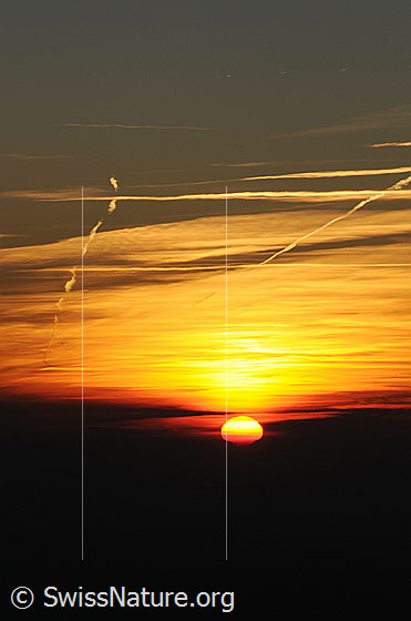 Foto: Sonnenuntergang. Die Sonne sinkt als helle Scheibe in eine Wolkenschicht. Der Himmel ist bewölkt und erscheint im Abendlicht in warmen Farbtönen zwischen rot und gelb.