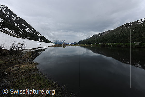 Foto: Regenwolken über dem Halsesee. Im ruhigen Bergsee spiegelt sich ein Bergrücken und am Ufer ist ein Schneefeld zu sehen.