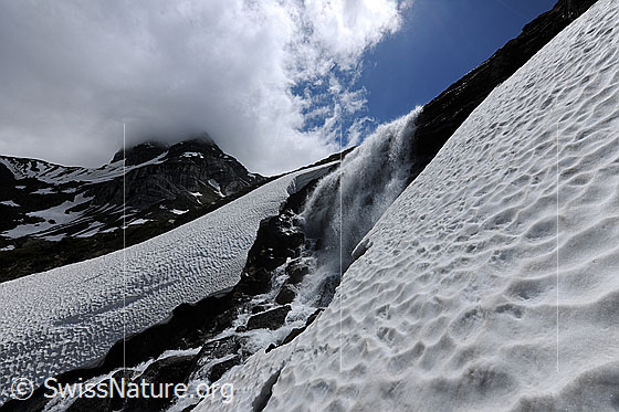 Foto: Schneereste und Schneefelder umgeben einen stiebenden Wasserfall, welcher über eine Felsstufe stürzt. Eine dunkle Quellwolke verhüllt die Gipfel im Hintergrund.