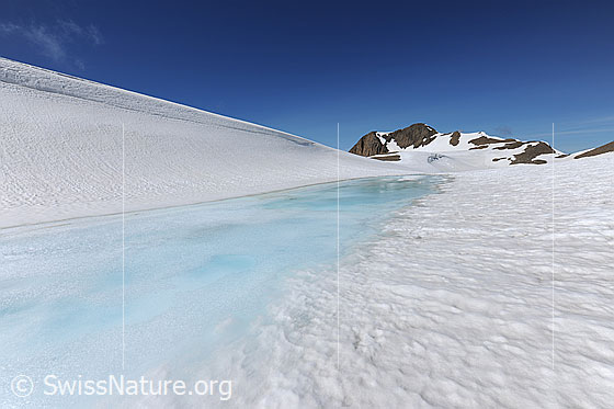 Foto: Schneebedeckte Hochebene mit zugefrorenem See und Berg im Hintergrund. Die Eisoberfläche ist angetaut und das Wasser über der Eisdecke erscheint in hellblauer Farbe.