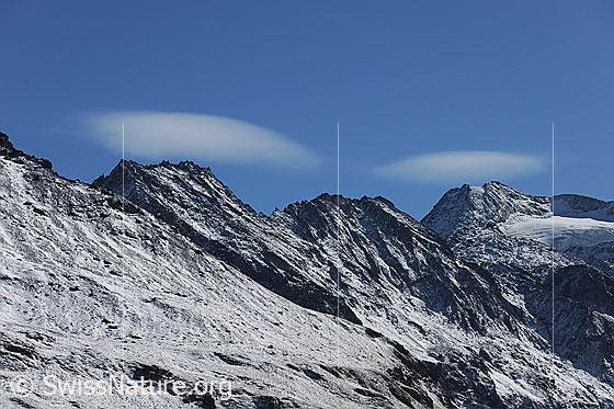 Foto: Aiguilles de la Lé und Pigne de la Lé. Über den leicht verschneiten Gipfeln zieren zwei Linsenwolken den blauen Himmel.