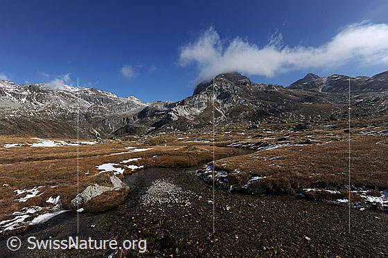 Foto: Urtümliche weite Berglandschaft im Herbst. Über die ausgedehnte Hochebene schlängelt sich ein Wasserlauf. Ein Wolkenband zieht sich über die Berggipfel.