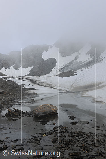 Foto: Geheimnisvolle Nebelstimmung in Berglandschaft mit Schneefeldern und seichtem Wasser am Ufer eines kleinen Bergsees.