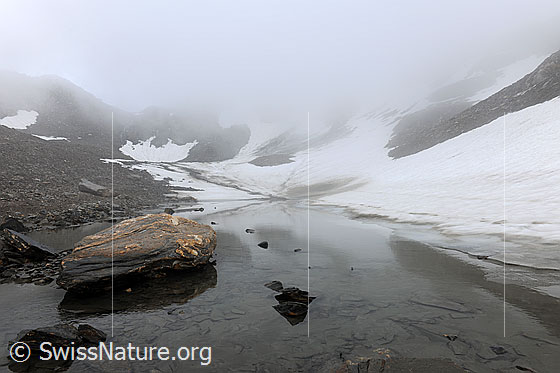 Foto: Mystische Berglandschaft mit Felsblock in kleinem Bergsee und Schneefeldern am Ufer.