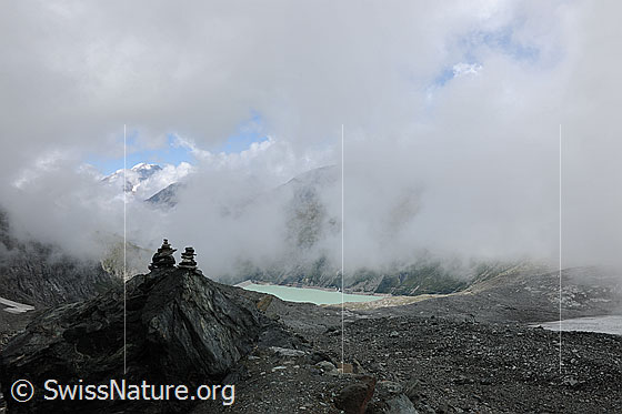 Foto: Weissmies in Wolkenlücke und Wolkenstimmung über dem Mattmark Stausee. Zwei Steinmännchen auf einem Felsblock markieren den Bergweg, welcher über die Seitenmoräne des Schwarzberggletschers führt.