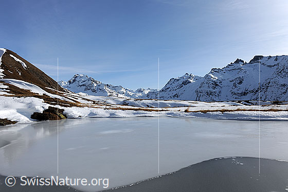Foto: Eisbedeckter Bergsee. Ausblick über die Eisfläche des Sees in eine Berglandschaft mit erstem Schnee im Herbst.