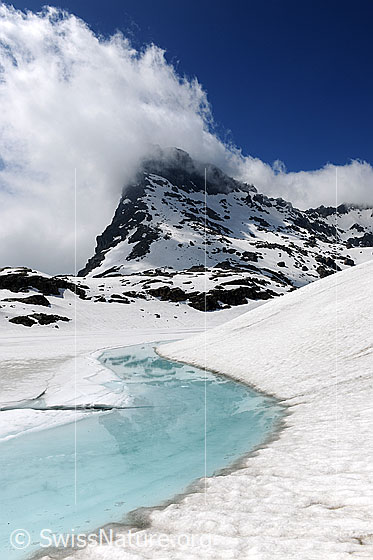 Foto: Hellblaues Wasser in tauendem Bergsee und Wolkenstimmung über der Berglandschaft. Am Ufer des Sees liegt Schnee.
